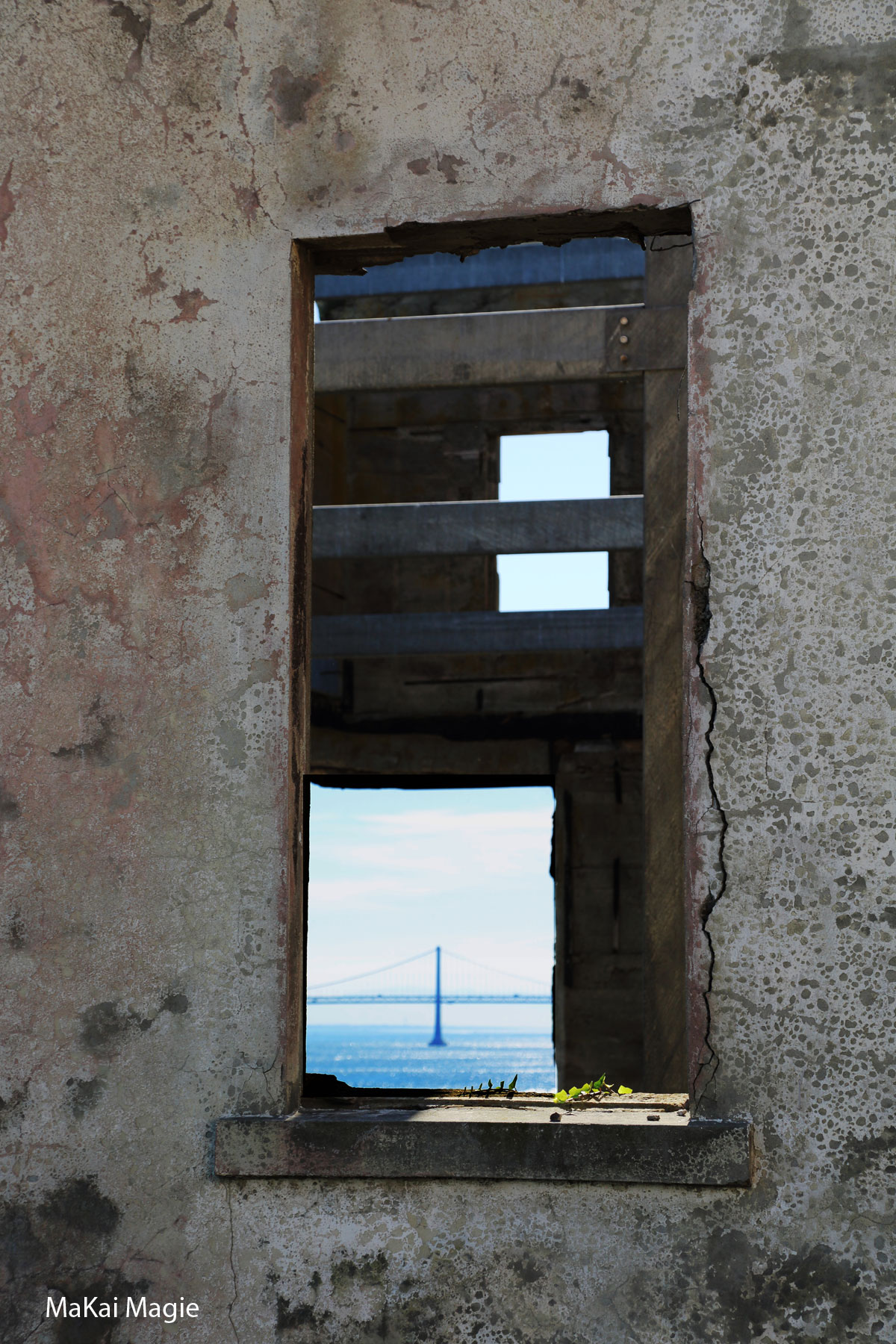 Photo of a narrow window in a worn stone wall, looking out at the San Francisco Bay Bridge above blue water.