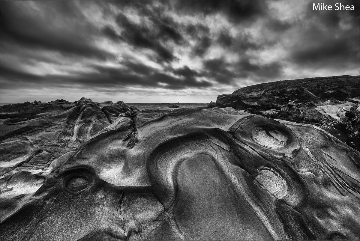 Photo of highly textured sandstone giving the impression of a horse head. Black and white, beach, ocean, and storm clouds in the background