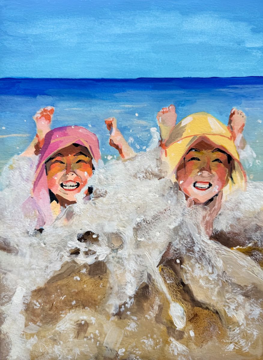 Two young girls, one with a pink hat and the other with a yellow hat, lay on their stomachs as shallow waves crash around them. They have big smiles on their faces.