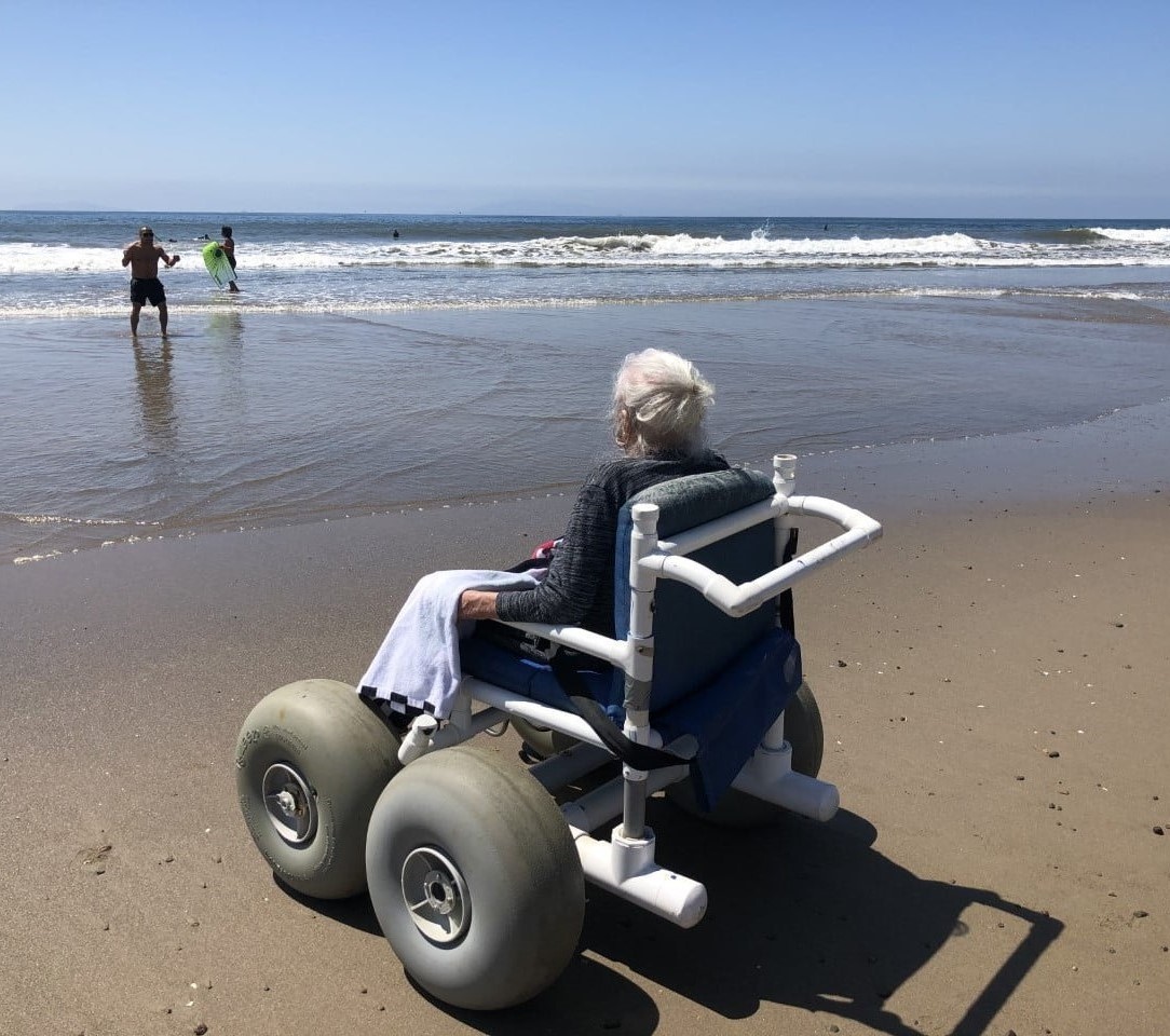 An older woman sits in a beach wheelchair, with large inflated wheels, on the beach watching family play in the surf