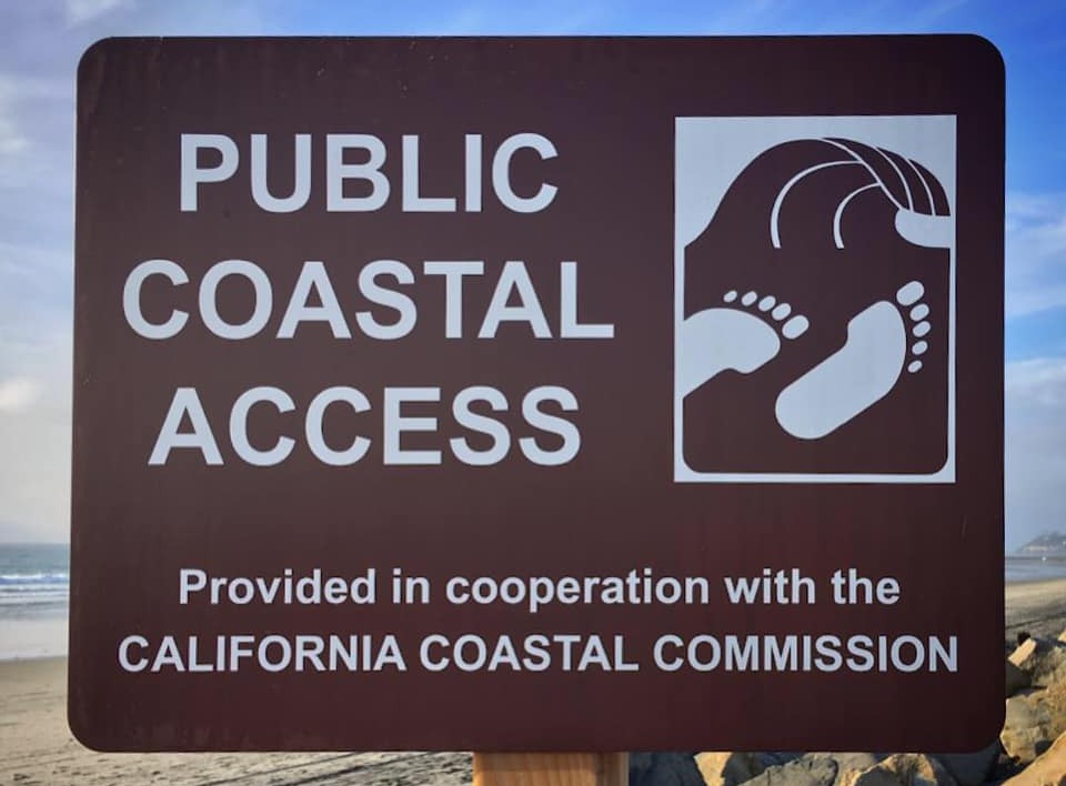 A brown sign next to a beach says Public Coastal Access provided in cooperation with the California Coastal Commission. The Coastal Access logo of two footprints below a wave is to the right of the text.