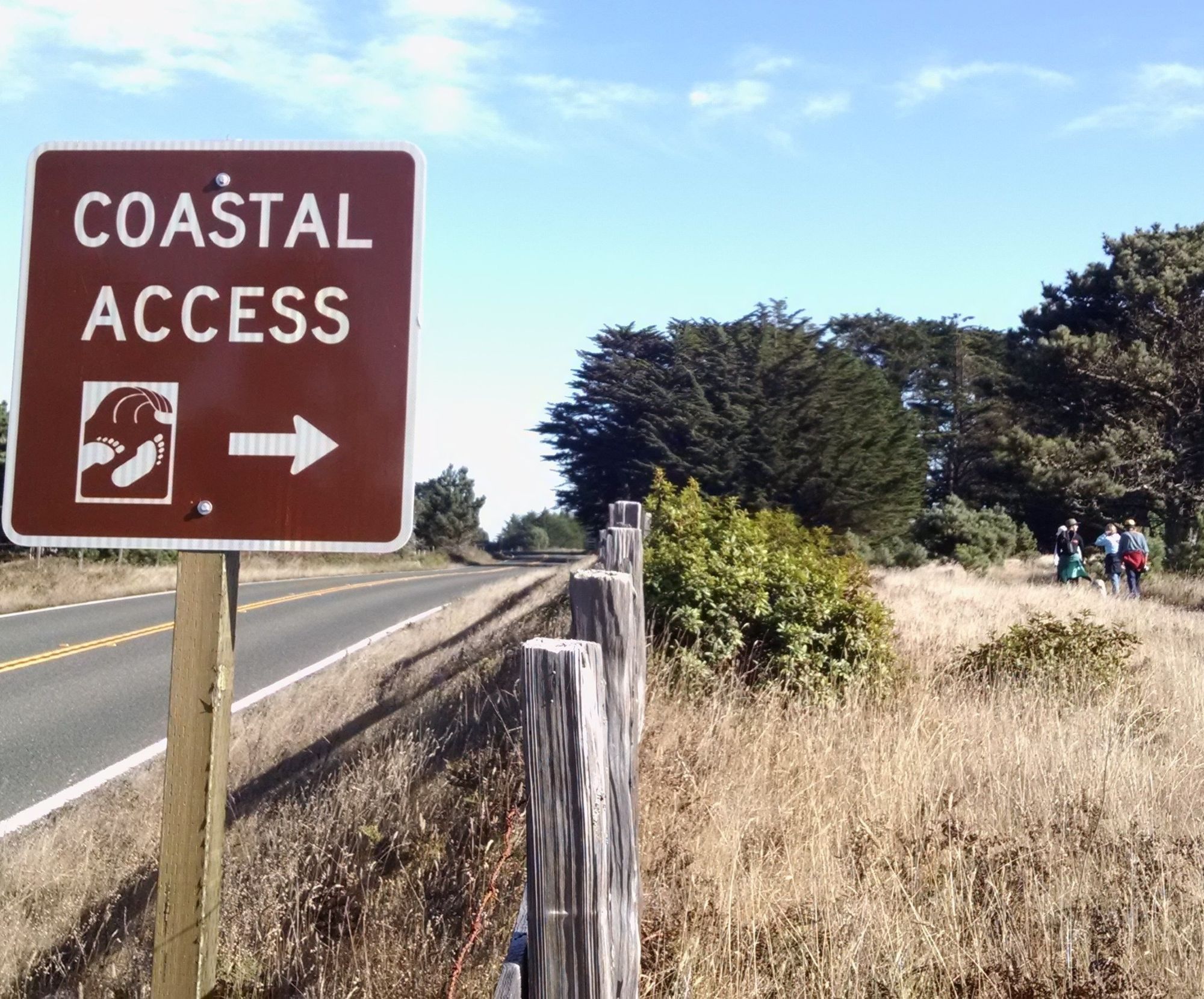 A coastal access sign stands next to a road as people walk along a coastal trail