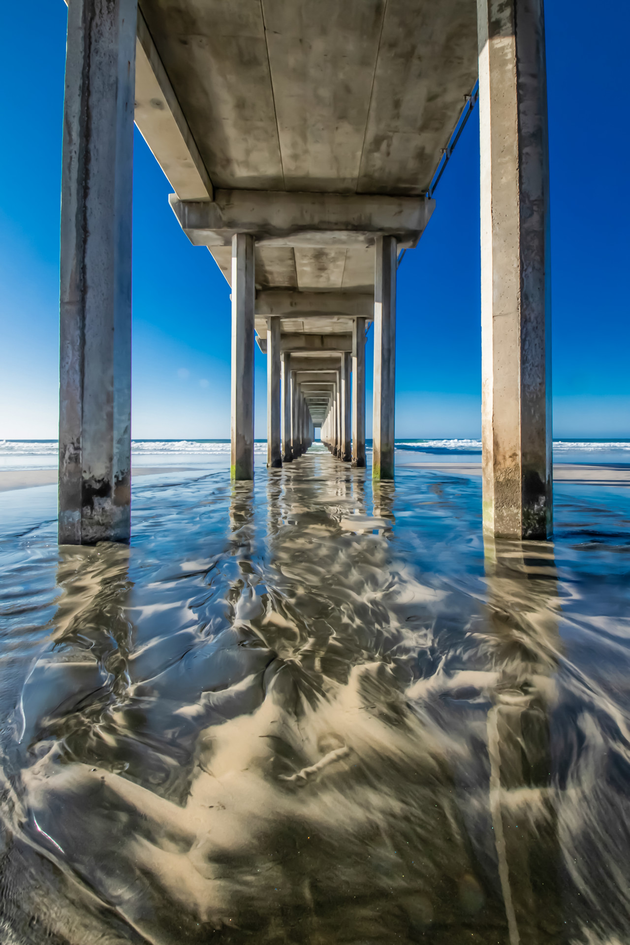 underneath the pier, looking down the length of the pier, water creates designs in the sand