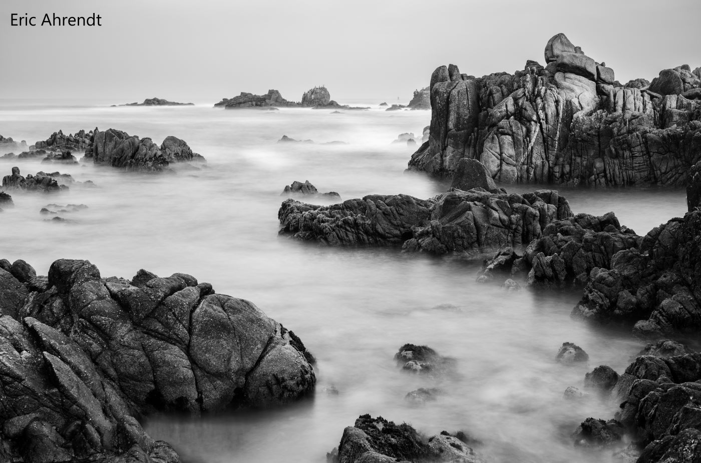 A black and white shows a moody scene of offshore rocks and misty waves