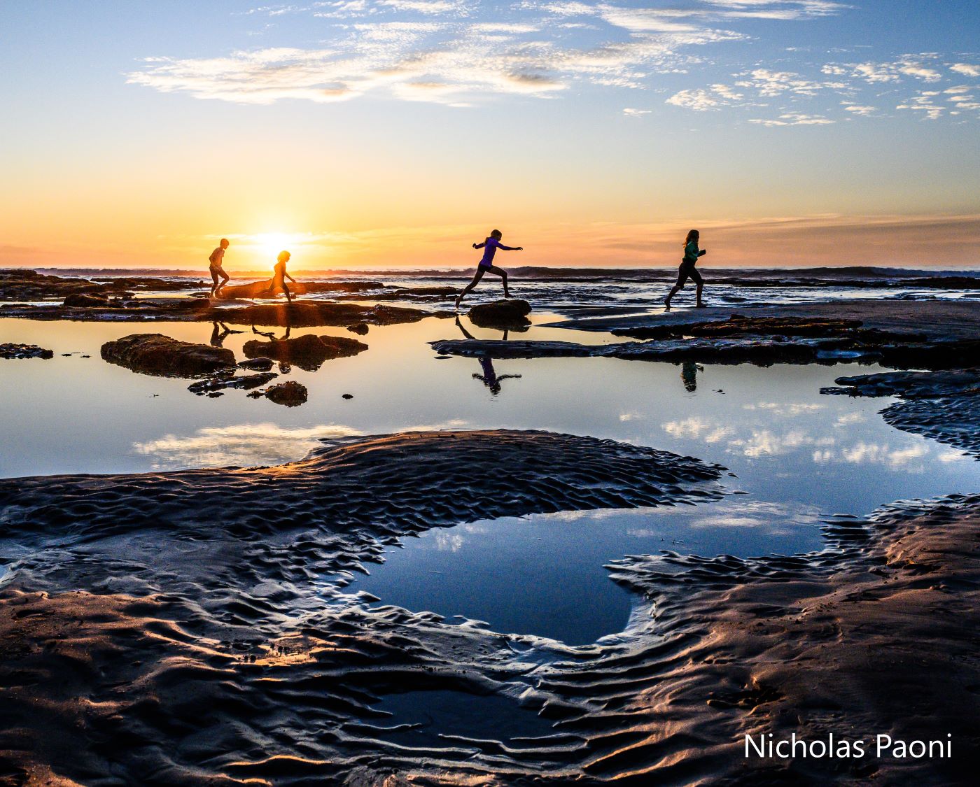 Four children are seen against an orange sunset as they leap across the rocks. The children and the sky is reflected in still pools of water in the foreground.