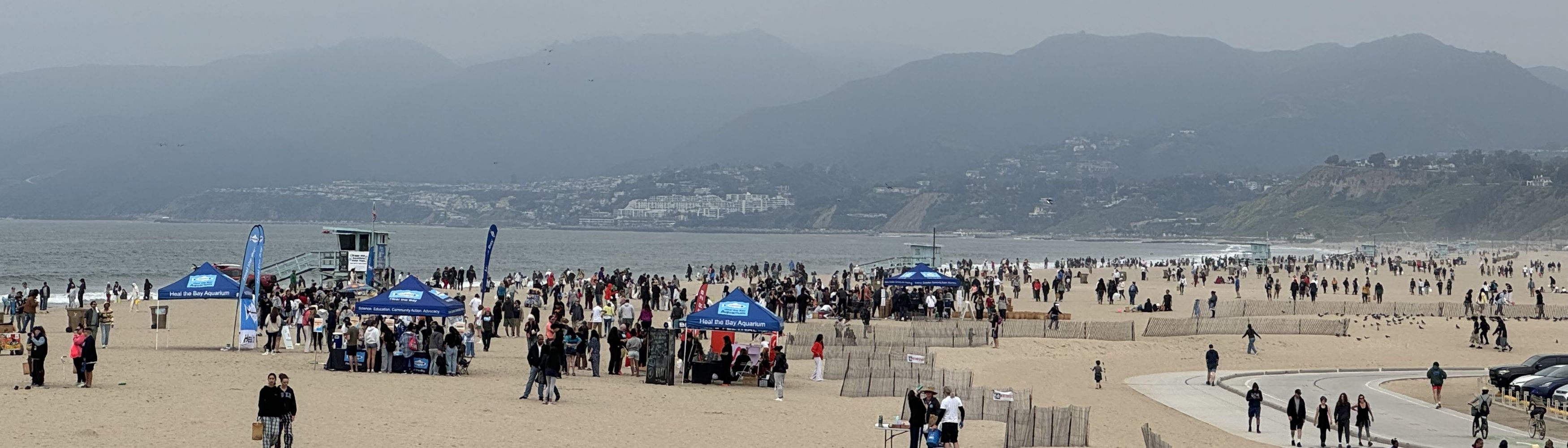 A crowd of people are participating in a beach cleanup on Santa Monica Beach. There are canopies on the sand with Heal the Bay on them.