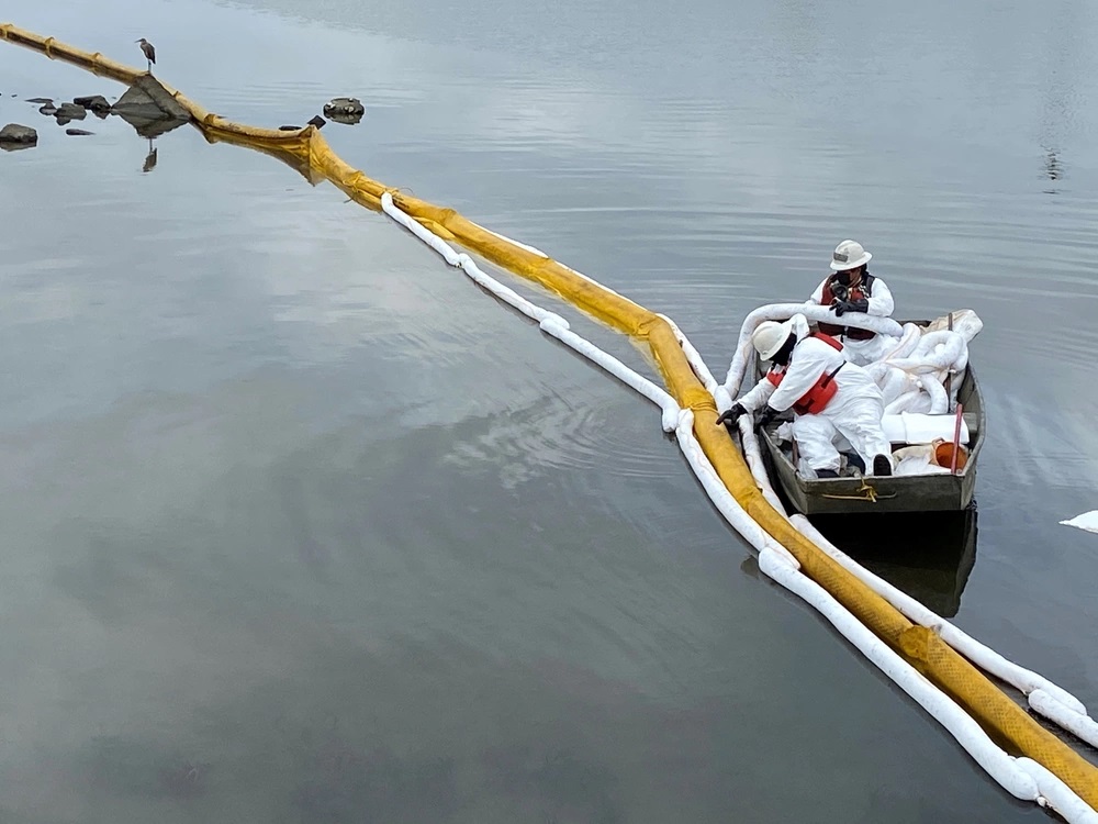 In a small boat, two people in protective gear adjust a boom on the water