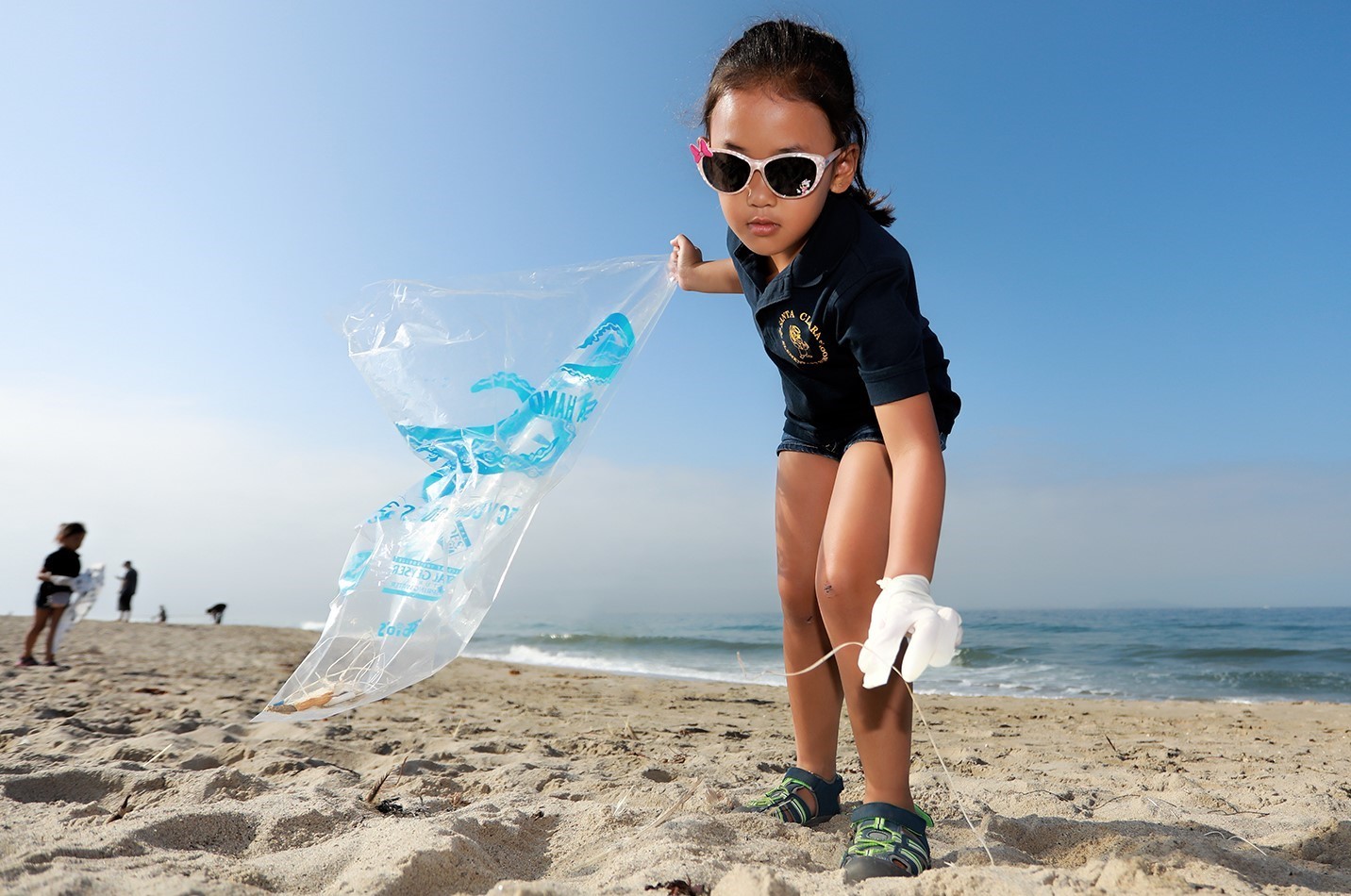 On a beach, a young child holds a trash bag in one hand as she leans down to pick up some fishing line