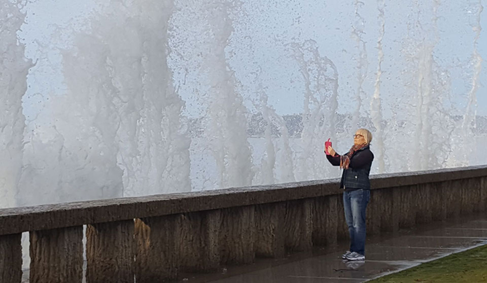 Photo of a woman photographing waves splashing up on the sidewalk at Butterfly Beach during king tides