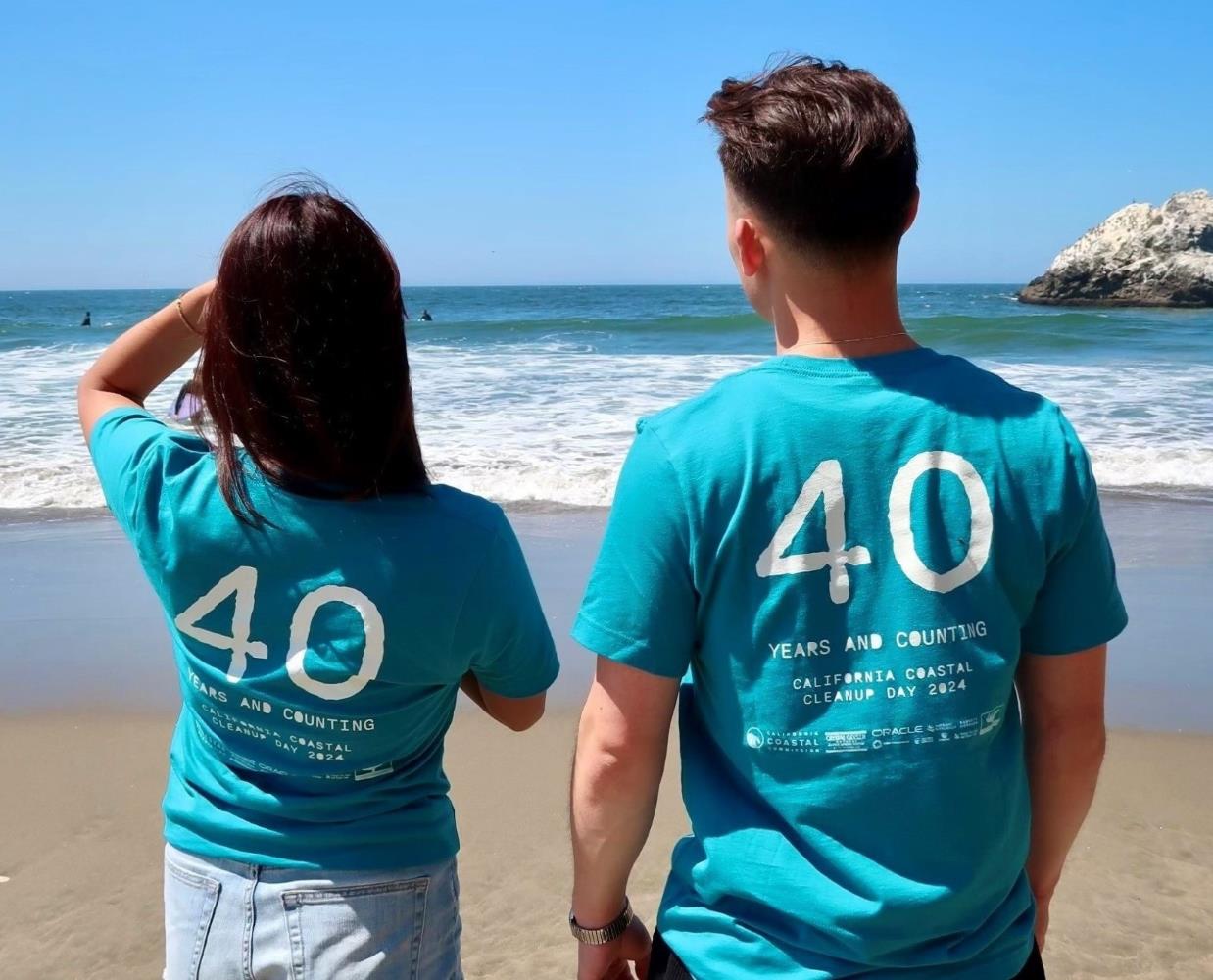 Two people are standing on the beach facing the ocean, showing the 2024 Coastal Cleanup Day back of shirt. 40 Years and Counting.