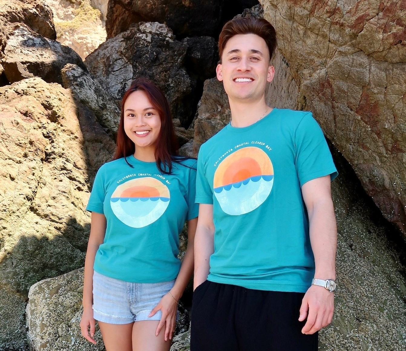 Two people standing in front of beach rocks wearing the 2024 Coastal Cleanup Day shirt showing the front graphic with round image of sun setting on the ocean, with teal background