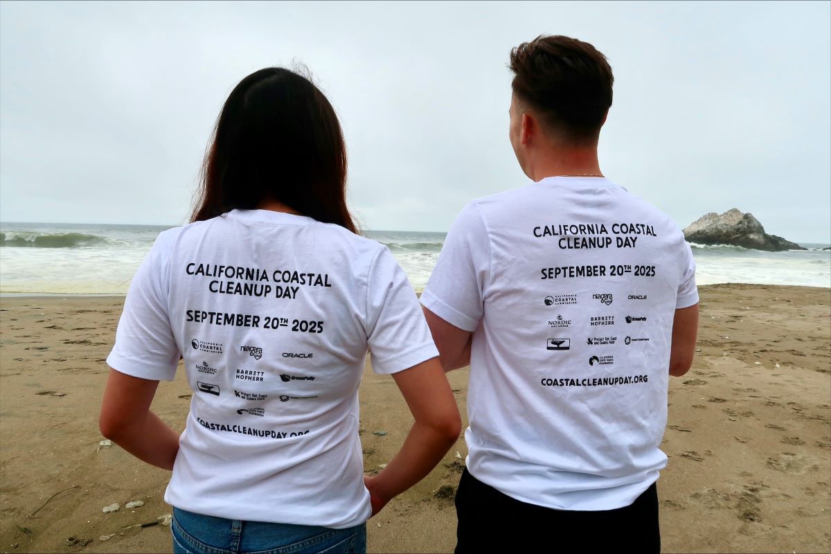 Two people are standing on the beach facing the ocean, showing the 2025 Coastal Cleanup Day back of shirt.