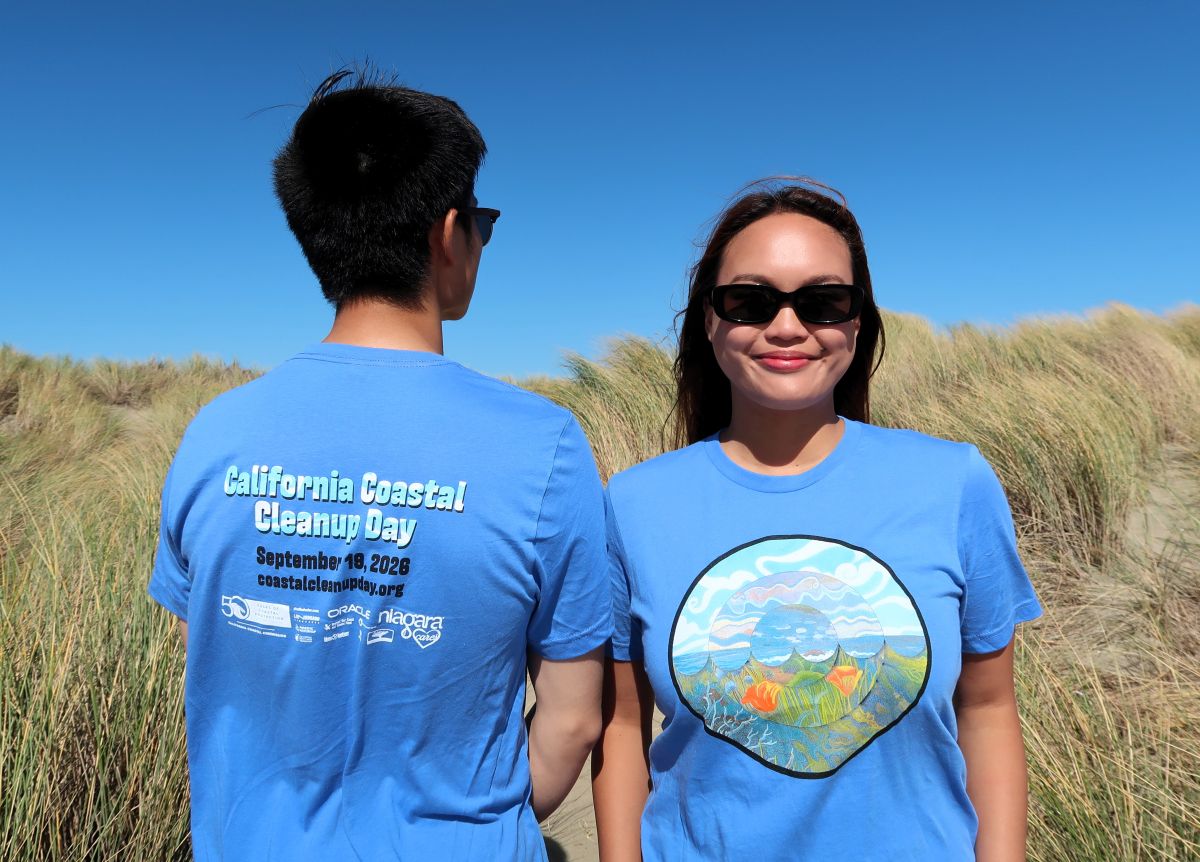 Two people standing in sand dunes wearing the 2026 
									Coastal Cleanup Day shirt. Shirt is light blue and has a round design on the front with the sky, ocean, and 
									California poppies. On the back is the event date, September 19, 2026, and sponsor logos.