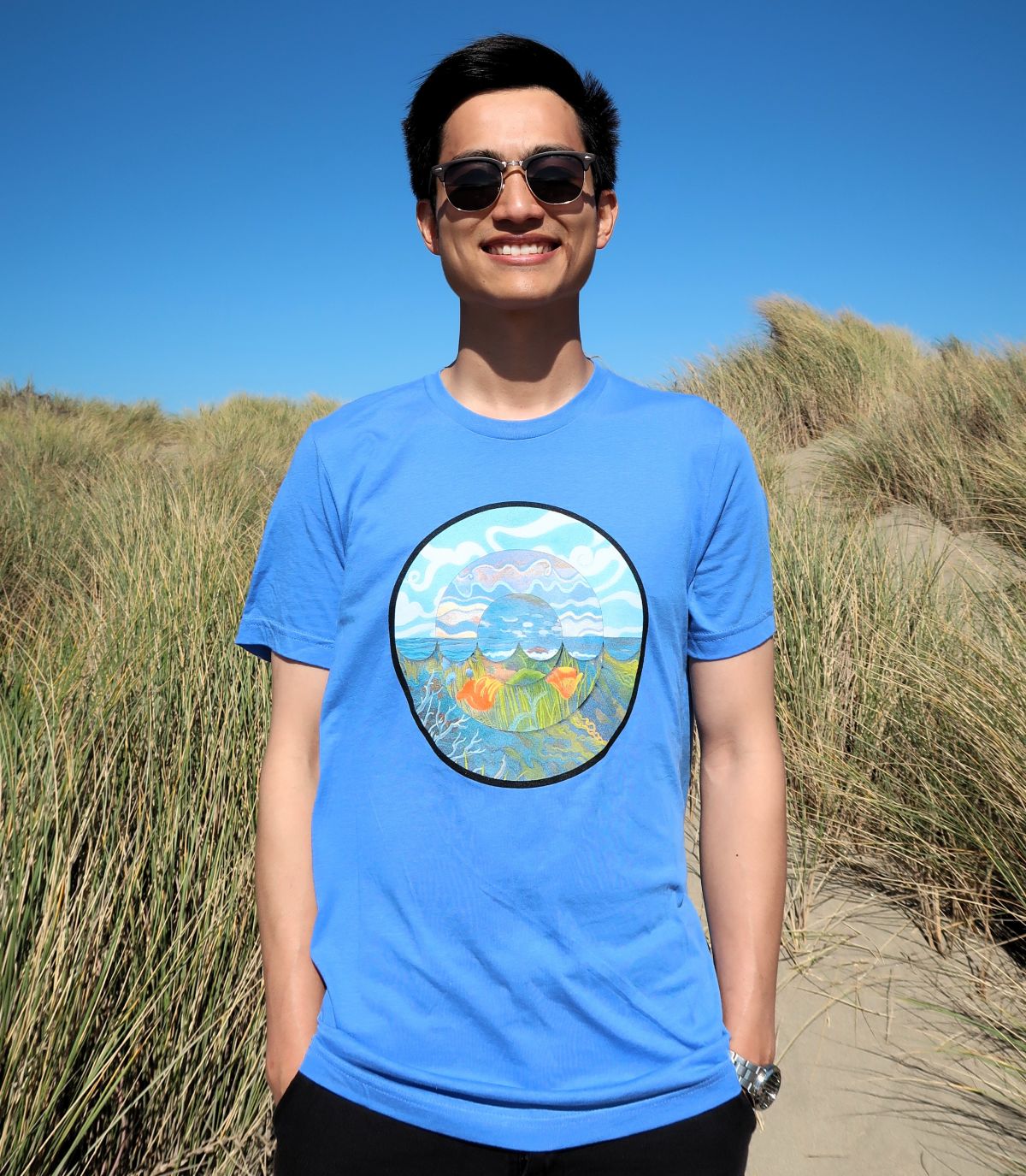 A man stands in sand dunes wearing the 2026 
									Coastal Cleanup Day shirt. Shirt is light blue and has a round design on the front with the sky, ocean, and 
									California poppies.