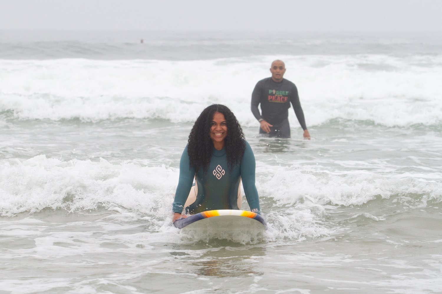 A man wades in the waves in the background while a woman in the foreground rides a surfboard with a smile on her face