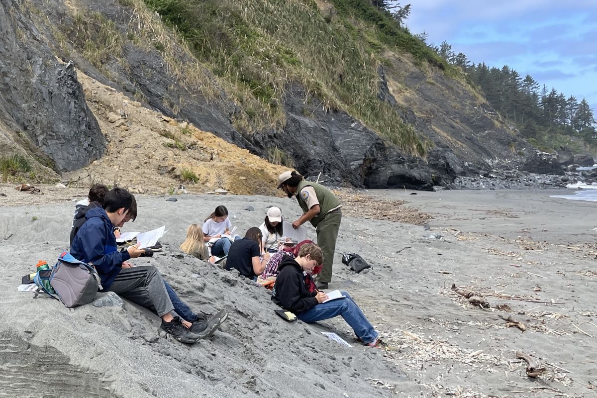A group of 7th grade students sit on a sandy beach to journal