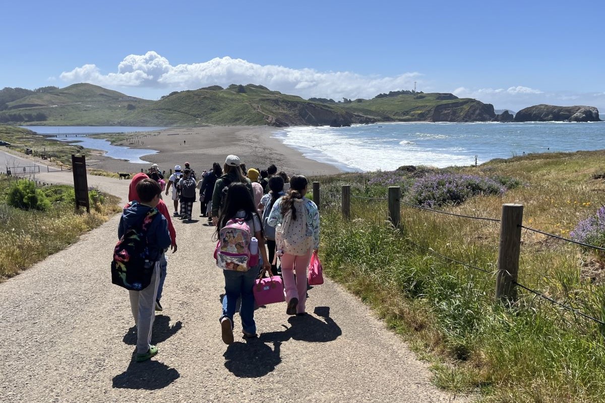 A large group of children wearing backpacks walk down a paved trail 
								toward a sandy beach with the ocean on the right and a lagoon on the left. 
								Green hills are on the other side of the beach. The sky is blue and sunny with 
								fluffy white clouds above the hills.