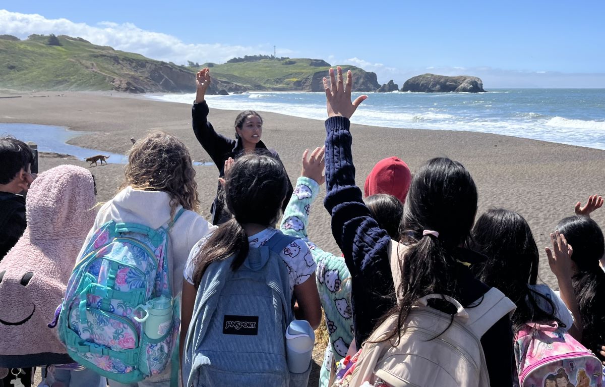 An instructor stands in front of a group of children as they raise their hands. They 
are standing on a beach on a sunny day.