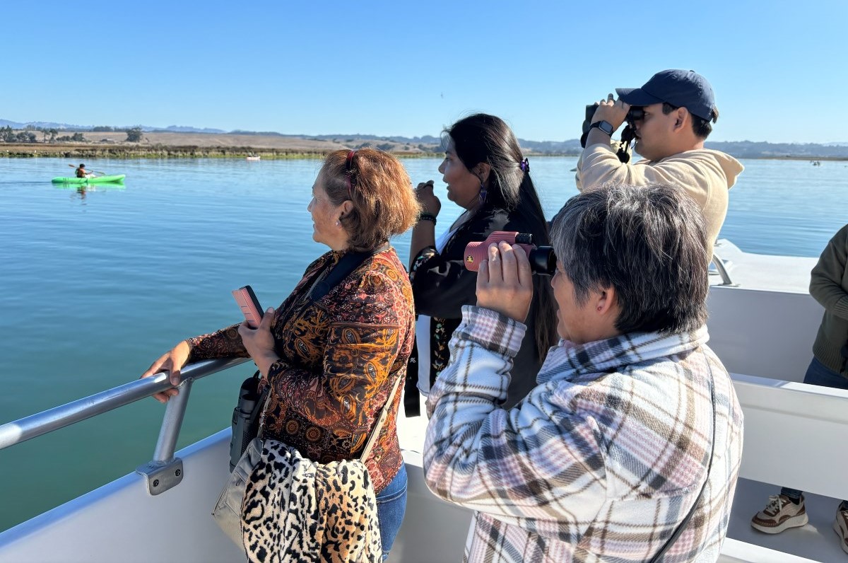 Four multi-generational Latino family members use binoculars to look at wildlife from 
								a boat in Elkhorn Slough. A kayaker is seen in the background.