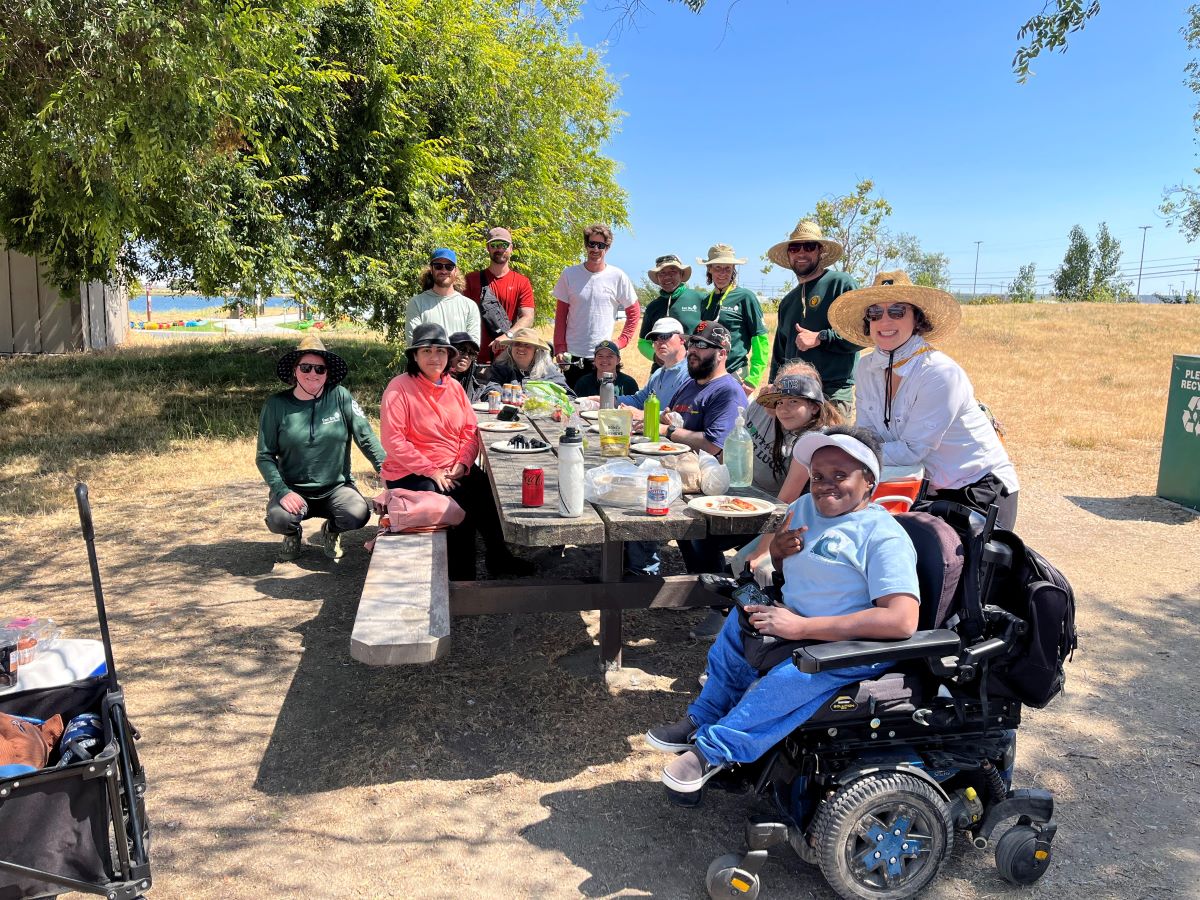 A group of about 15 adults, including one person in a wheelchaire, are gathered 
	around a picnic table enjoying a meal