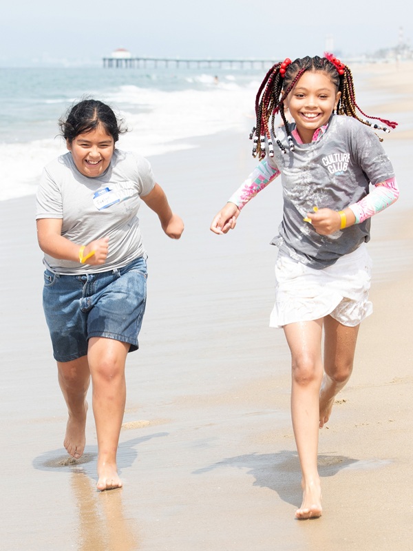 Two young people run down the sandy beach facing the camera with big smiles and windswept hair.