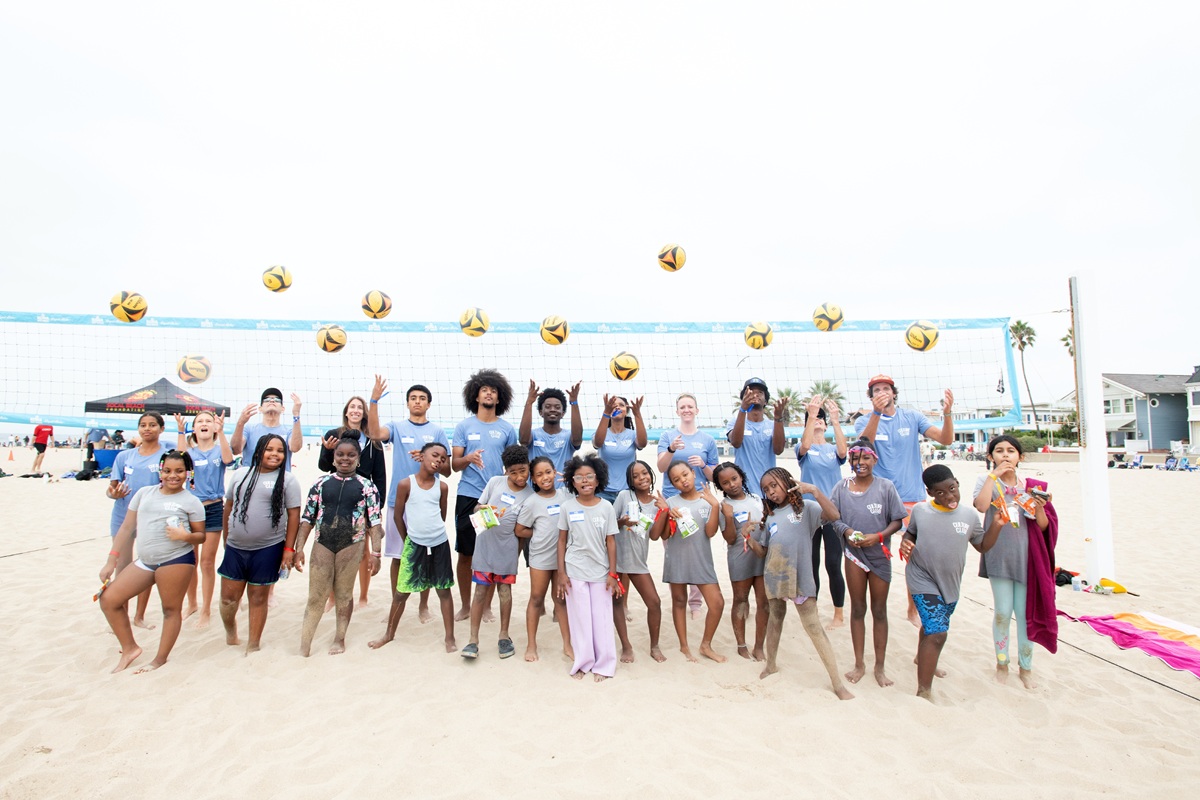 Twenty-six youth and volunteers pose in front of a beach volleyball net throwing beach volleyballs into the air and making silly faces.
