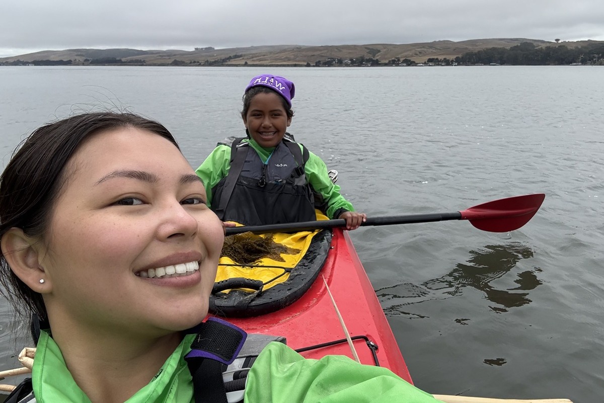 An adult and a youth sit in a kayak and smile at the camera with dark water reflecting the cloudy skies above. Rolling, yellow hills and trees
							sit at a distance on the horizon.