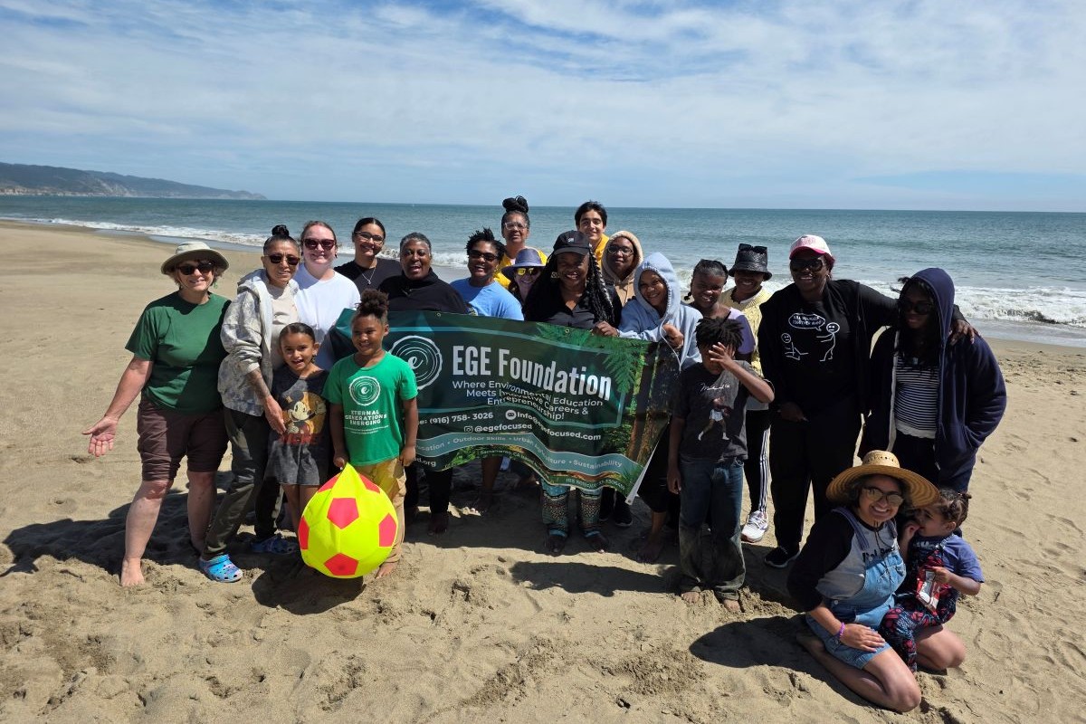 A group of 21 adults and youth stand on a sandy Point Reyes beach in front of blue skies and ocean waves. People in the center hold up an EGE 
				Foundation banner.